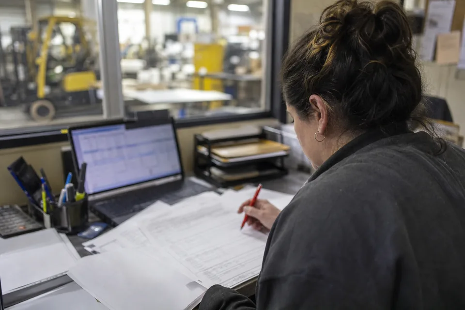 Procurement controller reviewing printed purchase orders at a plant office desk with the factory floor behind